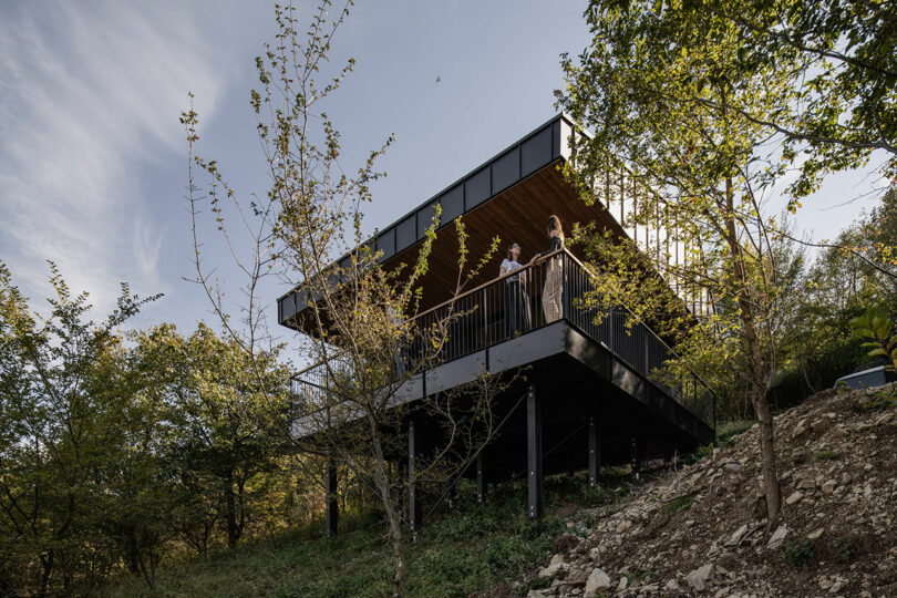 Modern elevated cabin with metal supports and a large balcony, situated on a sloped, wooded hillside. Several people stand on the balcony looking out over the landscape.