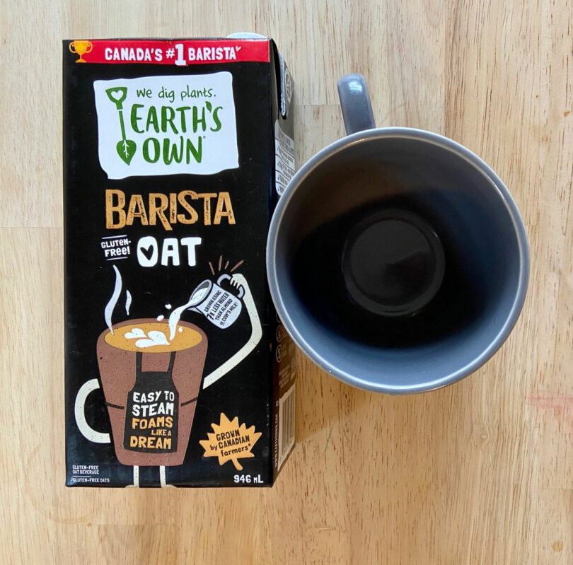 A carton of Earth's Own Barista Oat milk, captured in a Katherine Duclos-inspired style, sits next to an empty black mug on a wooden surface.