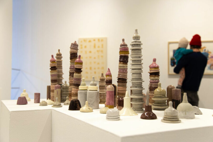 Ceramic sculptures resembling small towers by Katherine Duclos are displayed on a white pedestal in an art gallery; a person holding a child views artwork in the background.