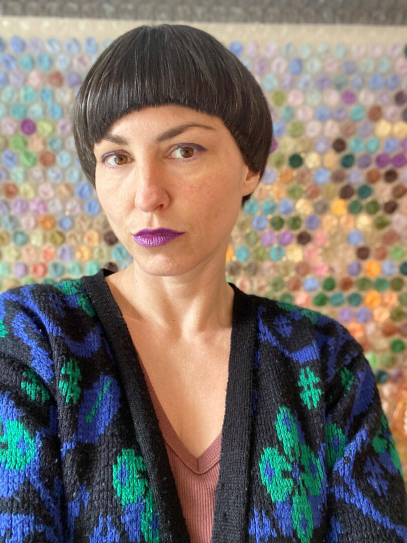 Katherine Duclos, with short dark hair and purple lipstick, poses in front of a colorful, circular-patterned background, wearing a blue and black patterned sweater over a pink top.