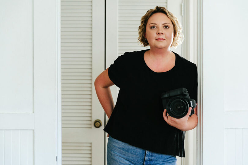 A person with short curly hair, wearing a black shirt and jeans, stands indoors holding a professional camera.