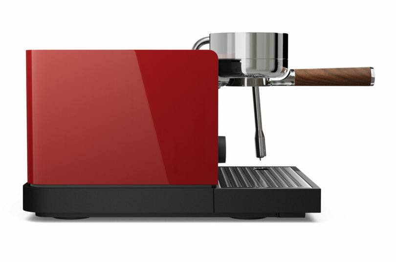 Side view of a modern, red Fellow espresso machine with a metallic portafilter and a wooden handle, set against a white background.