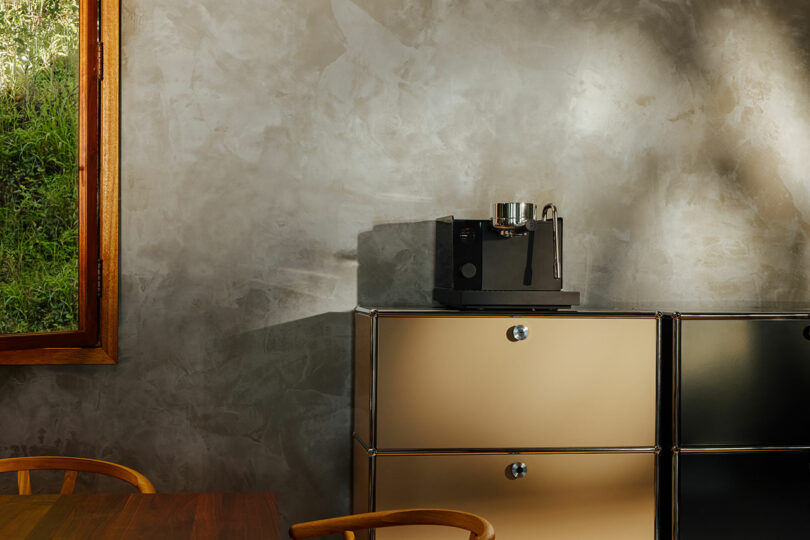 A black Fellow espresso machine sits on top of beige metal cabinets next to a window in a room with concrete walls and wooden furniture.