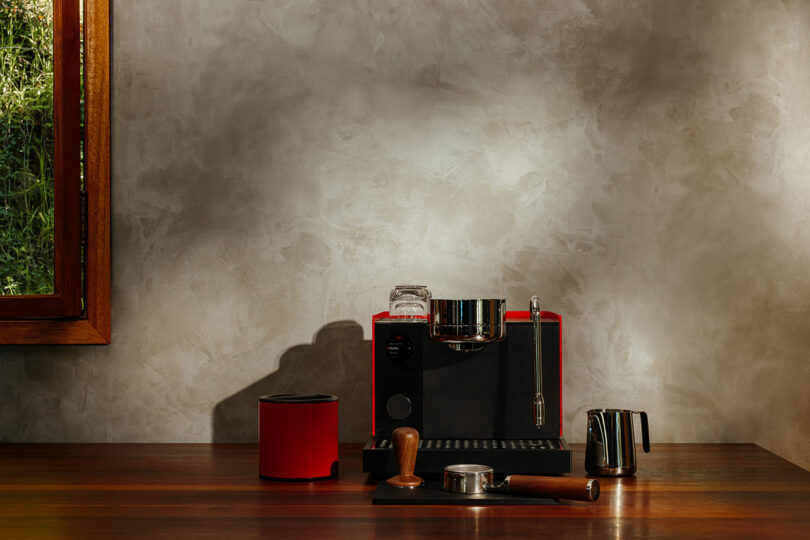 A red espresso machine with Fellow coffee-making tools and a milk frothing pitcher sits on a wooden counter against a textured wall, sunlight streaming through the window.