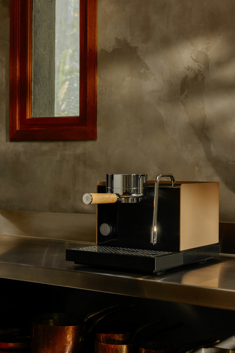A modern Fellow espresso machine with a wooden handle sits on a stainless steel countertop beneath a window with a red frame, against a textured gray wall.