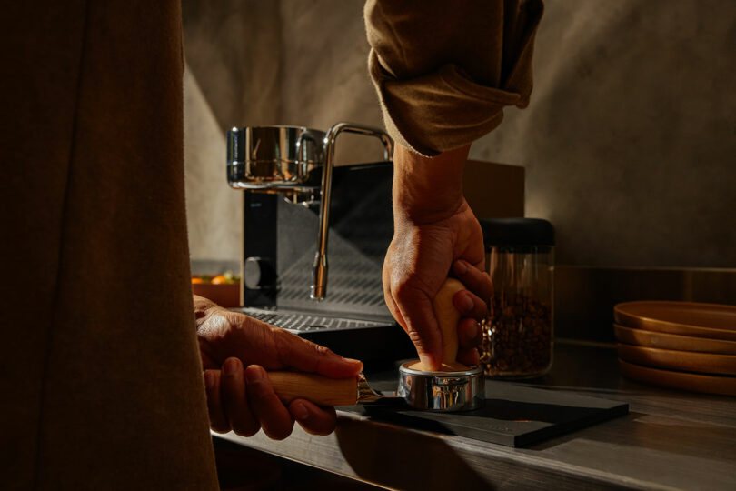 A person uses a Fellow tamper to press coffee grounds into a portafilter beside a black espresso machine on a countertop.