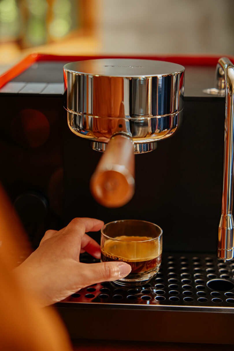 A person holds a Fellow glass cup under the portafilter of an espresso machine, collecting freshly brewed coffee.