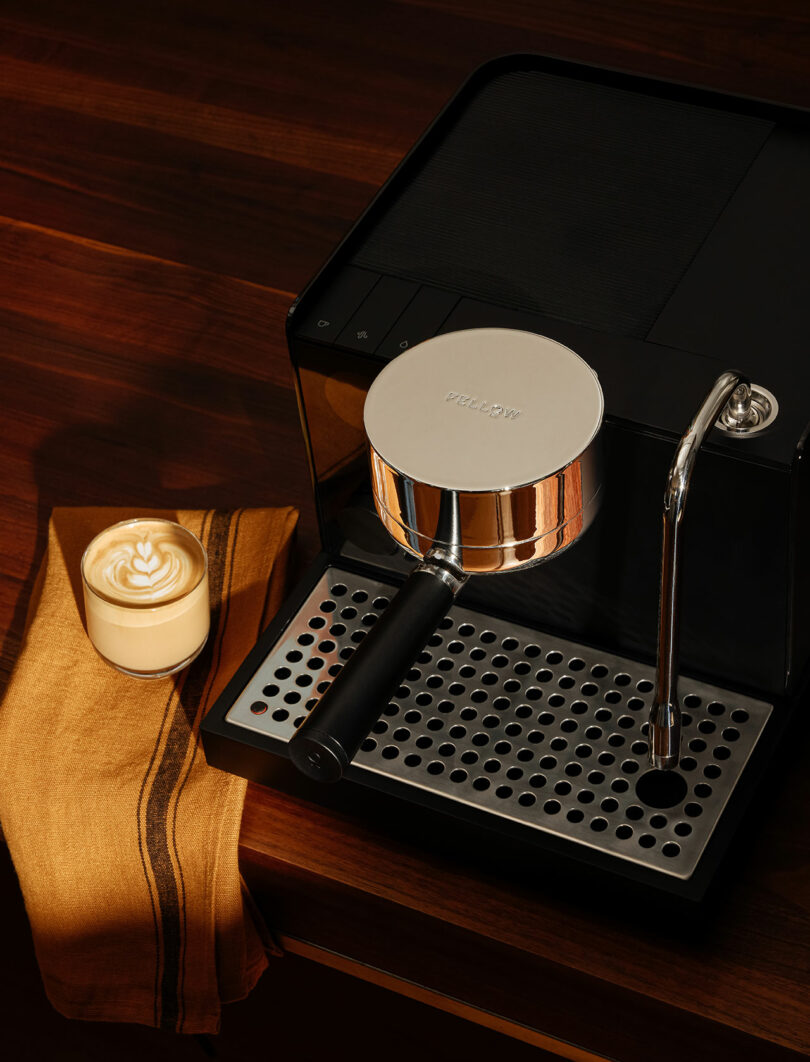 A sleek black Fellow espresso machine sits on a wooden counter next to a brown-striped towel and a glass of latte with latte art on top.