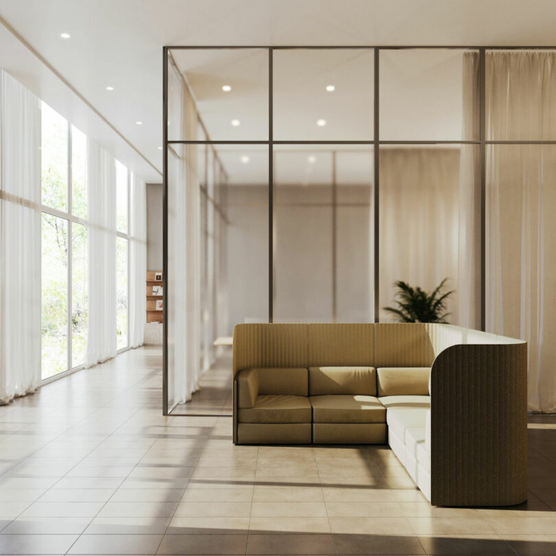 Modern office lounge with a beige sectional sofa, glass partition walls, tile floor, large windows, and natural light. A potted plant is visible in the background.