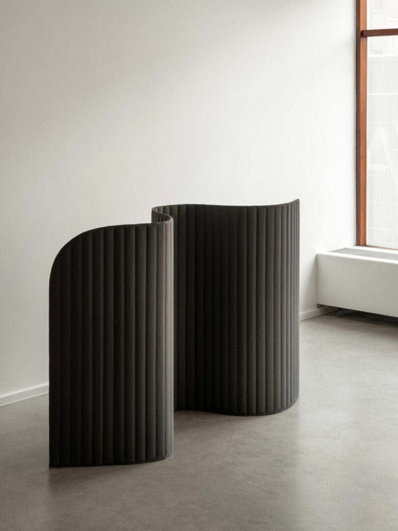 A black, wavy, ribbed room divider stands on a gray floor near a white wall and large window in a minimal, modern interior.