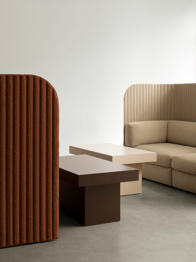 Minimalist interior with neutral-toned modular sofa, two rectangular coffee tables, and a textured brown room divider on a gray floor against a plain white wall.