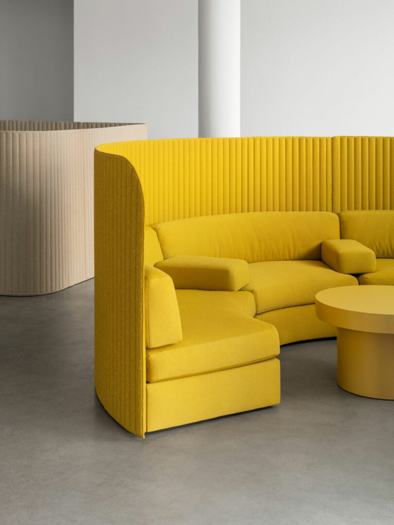 Curved yellow sectional sofa with high ribbed backrest and matching round coffee table in a minimalist, neutral-toned room with concrete floor.