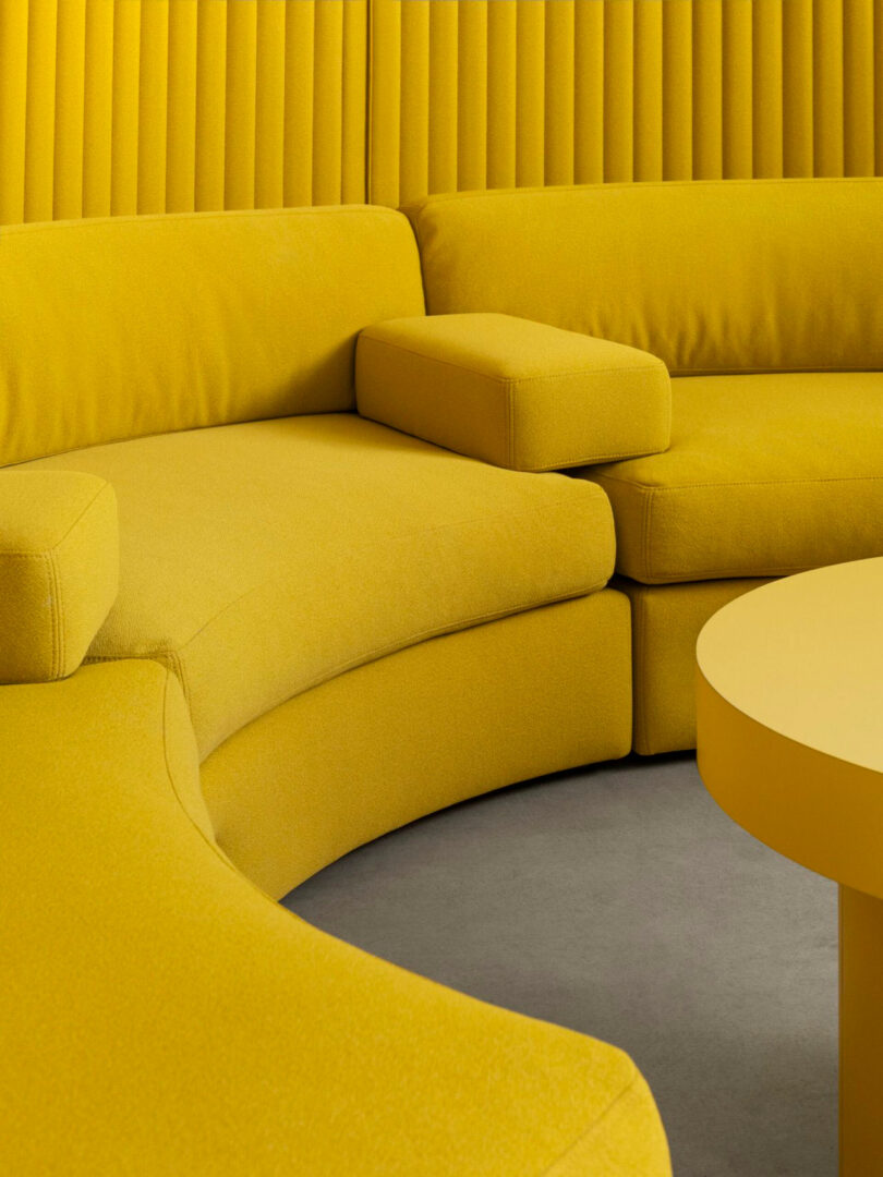 Curved yellow sofa with matching armrest and round yellow table on grey floor, set against a yellow wall with vertical paneling.