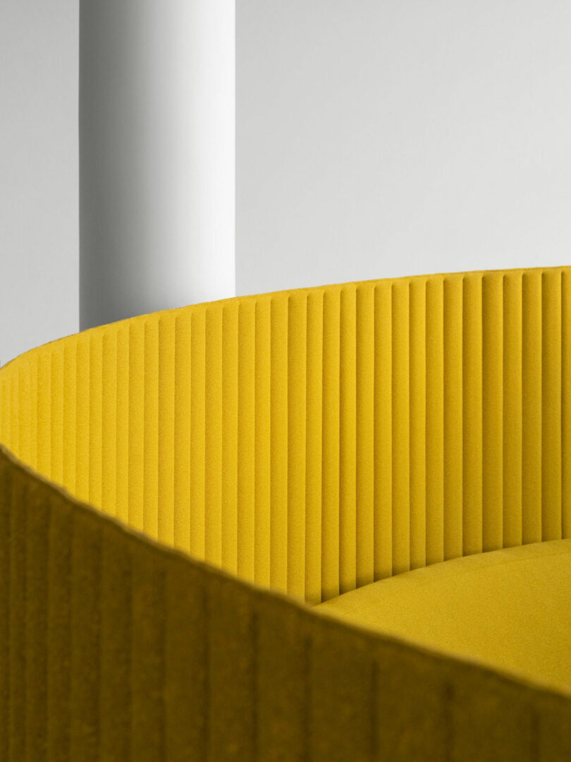 Close-up of a modern yellow upholstered chair with vertical ridges, positioned next to a white cylindrical column against a plain light background.