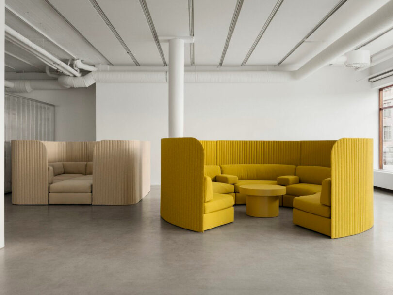 Modern office lounge with yellow and beige modular seating areas, round tables, and exposed white ceiling pipes in a bright, minimalist space.