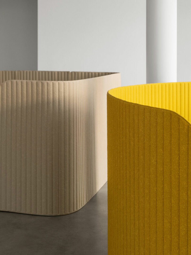 Curved beige and yellow ribbed partitions stand on a gray floor in a minimalist room with white walls and a column.