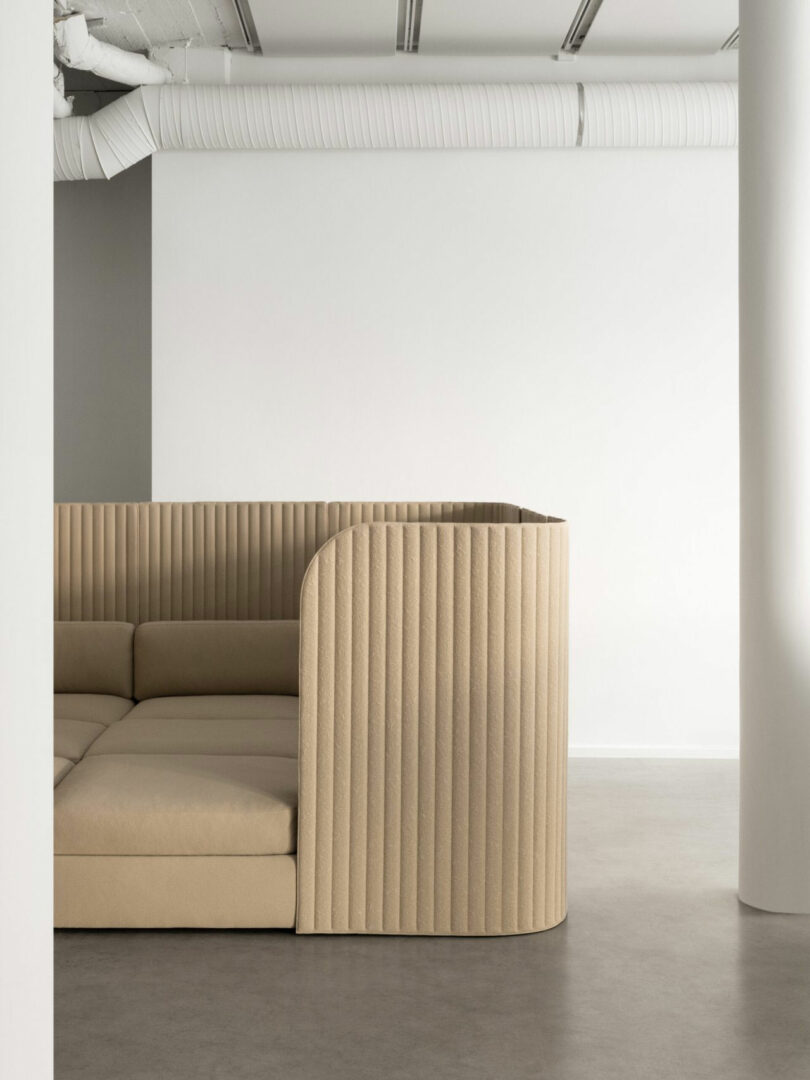 A modern beige sectional sofa with ribbed upholstery is positioned in a minimalist room with white walls, exposed ductwork, and a concrete floor.