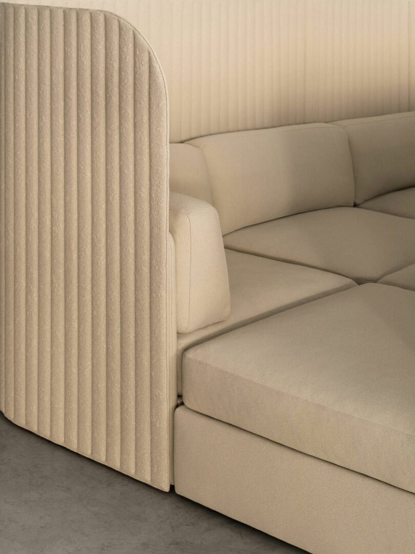 A close-up of a beige, modular sofa with ribbed upholstery on the side panel and plush, square cushions on a concrete floor.
