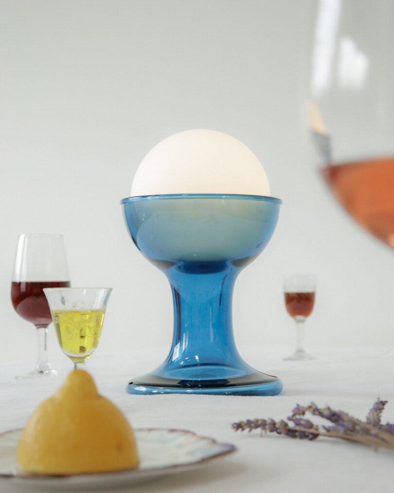 A blue glass lamp with a white globe sits on a table set with assorted drinks, a pear dessert, and lavender sprigs against a plain white background.