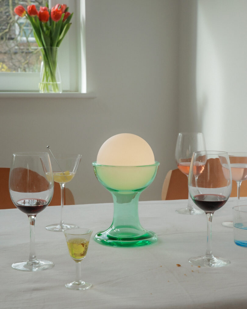 A table with several wine and cocktail glasses, a glowing round lamp in a green holder, and a vase of tulips on a windowsill in the background.