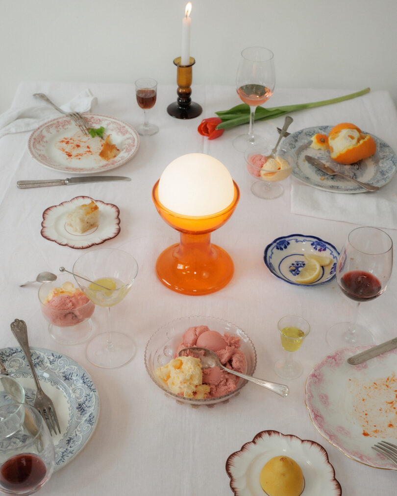 A table with partially eaten desserts, wine glasses, plates, candles, and a tulip on a white tablecloth. An orange lamp is lit at the center.