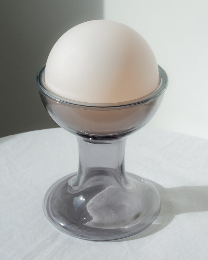 A white egg sits upright in a transparent glass egg cup on a white surface with a plain light background.