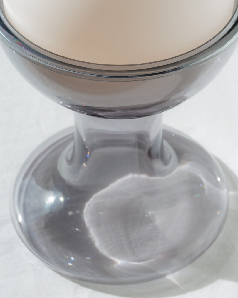 A close-up of a clear glass stand with a round, off-white object resting on top, casting a shadow and reflection on a white surface underneath.