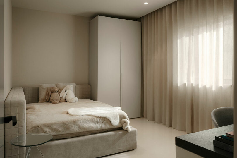 Minimalist bedroom with neutral tones featuring a bed with plush toys, a large window with beige curtains, a wardrobe, and a small side table.