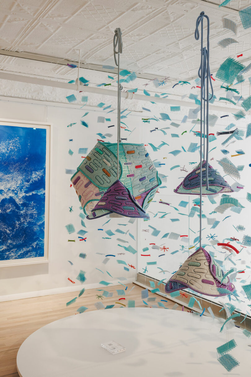 Three geometric, textile sculptures hang from the ceiling amid scattered mesh pieces in a brightly lit, modern gallery with a blue abstract artwork on the wall.