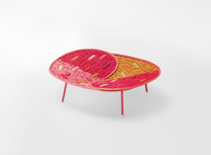 A colorful, oval-shaped bench with red legs and a woven seat featuring bright pink, red, and yellow patterns, set against a plain white background.
