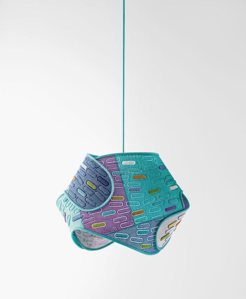 A geometric, multicolored pendant lamp with a textured surface hangs from a turquoise cord against a plain white background.