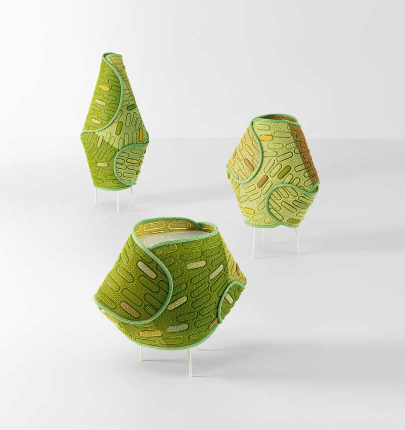 Three geometric fabric sculptures with green and yellow patterns stand on thin metal legs against a plain white background.