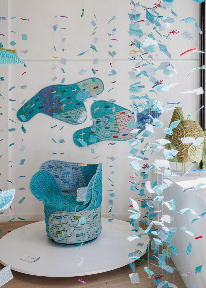 Blue and turquoise textile art installation with a patterned chair, wall hangings shaped like hands, and suspended fabric pieces in a bright gallery space.