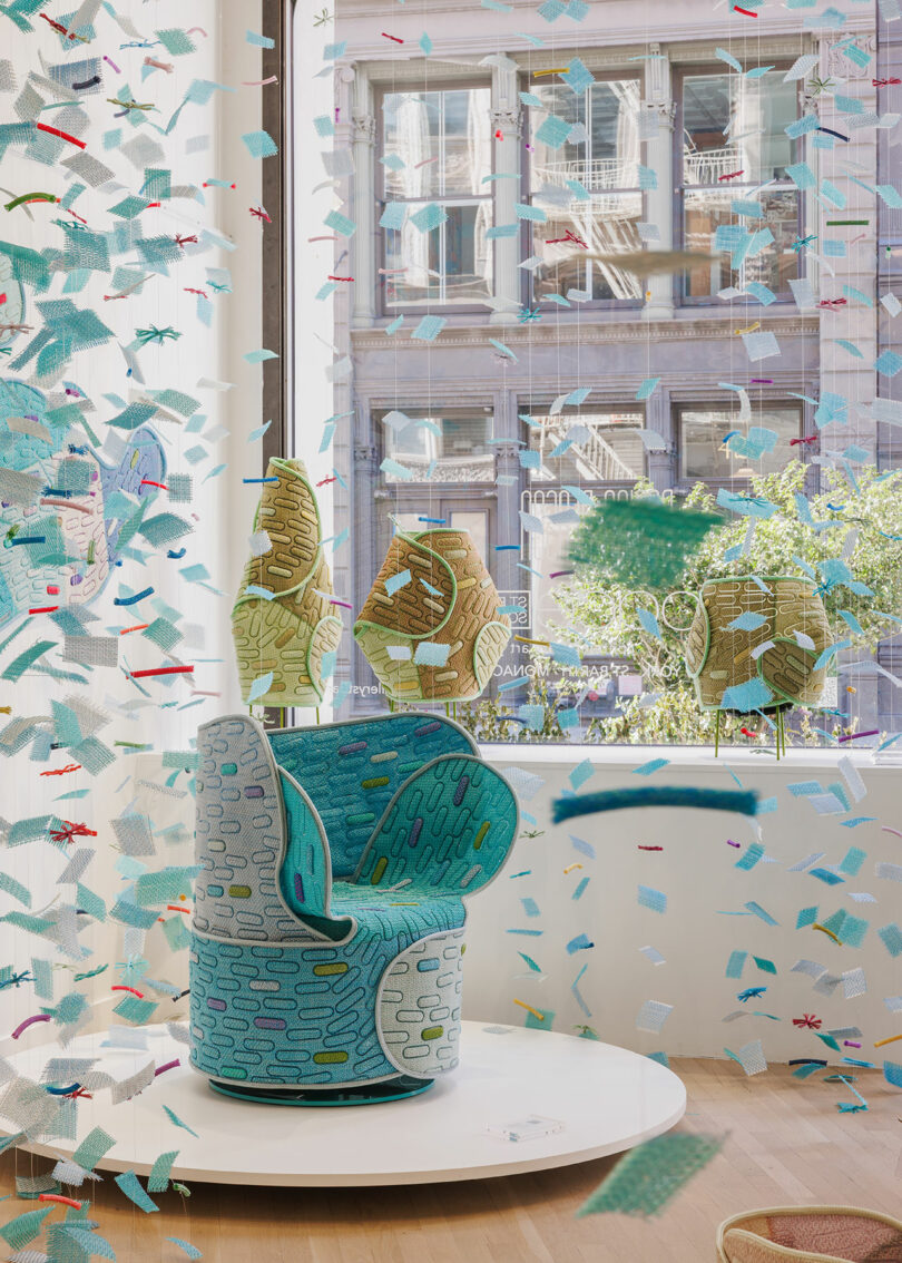 A patterned swivel chair sits on a white platform in front of a large window, surrounded by blue and teal confetti-like decorations, with buildings visible outside.