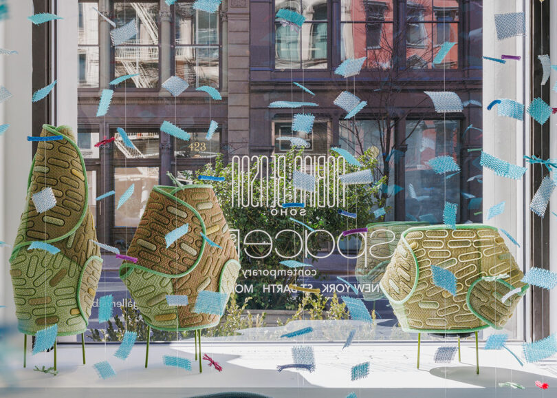Display window features three textured sculptural objects with abstract patterns, set against a street view and glass decorated with blue and white confetti-like shapes.