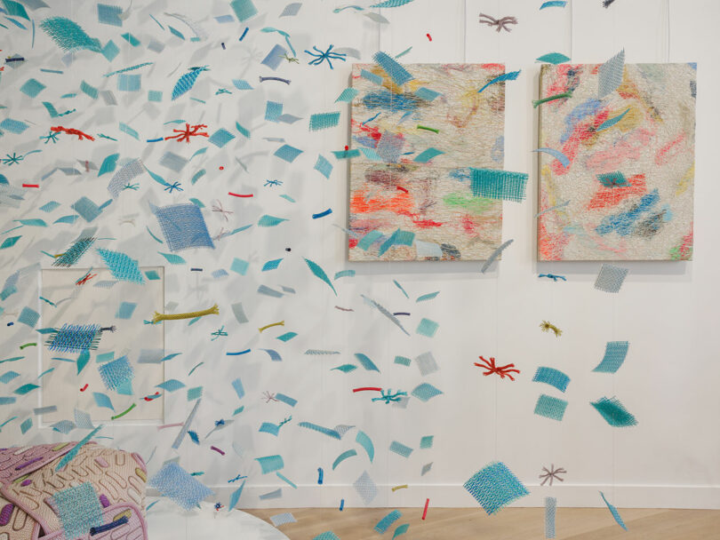 Mixed media art installation with blue mesh squares and colored thread pieces suspended in air, two textured canvas artworks on a white wall, and a patterned pillow in the lower left corner.
