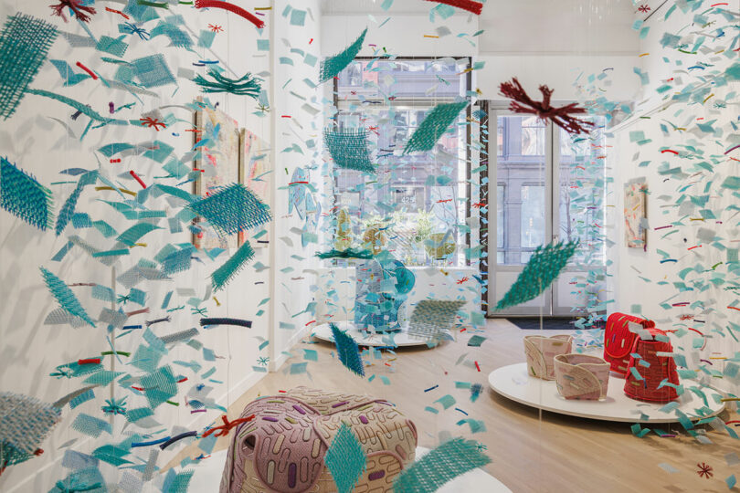 Colorful woven textile pieces are suspended in midair throughout a bright gallery space with modern seating and large windows.