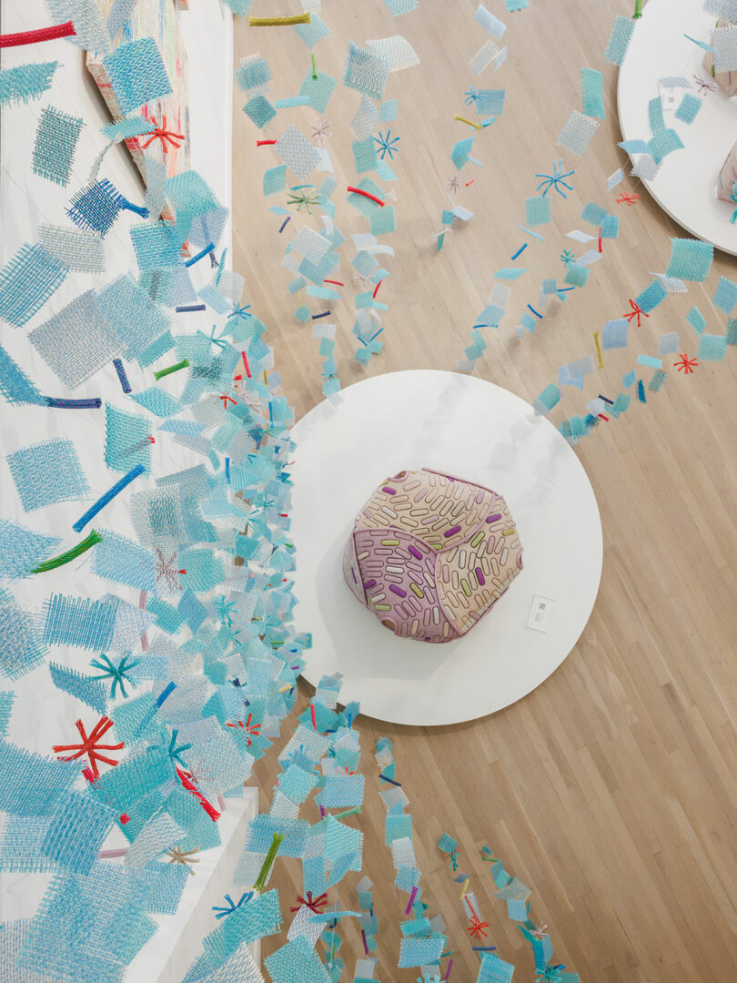 An overhead view of a round ottoman with a patterned cushion on a white platform, surrounded by blue textured decorations suspended in midair over a light wood floor.