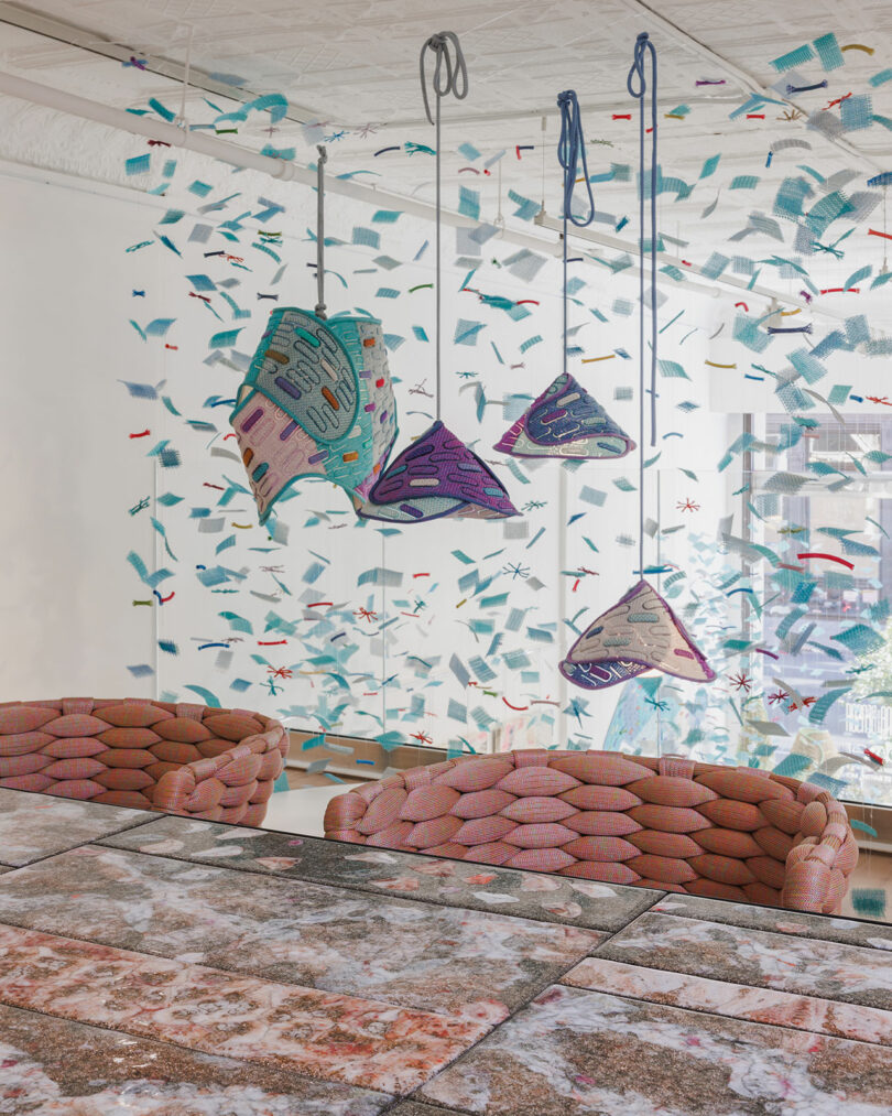 A dining area with pink woven chairs, a marble table, and four colorful geometric pendant lights; abstract confetti-like decorations fill the glass wall behind.