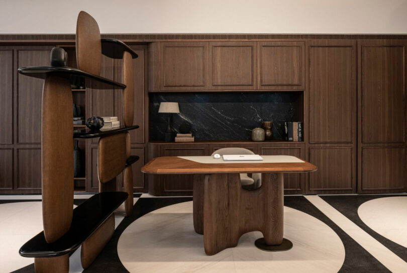 Modern office with dark wood furniture, a sculptural desk, built-in cabinets, open shelves, and a marble backsplash. Neutral color palette with black and white flooring.