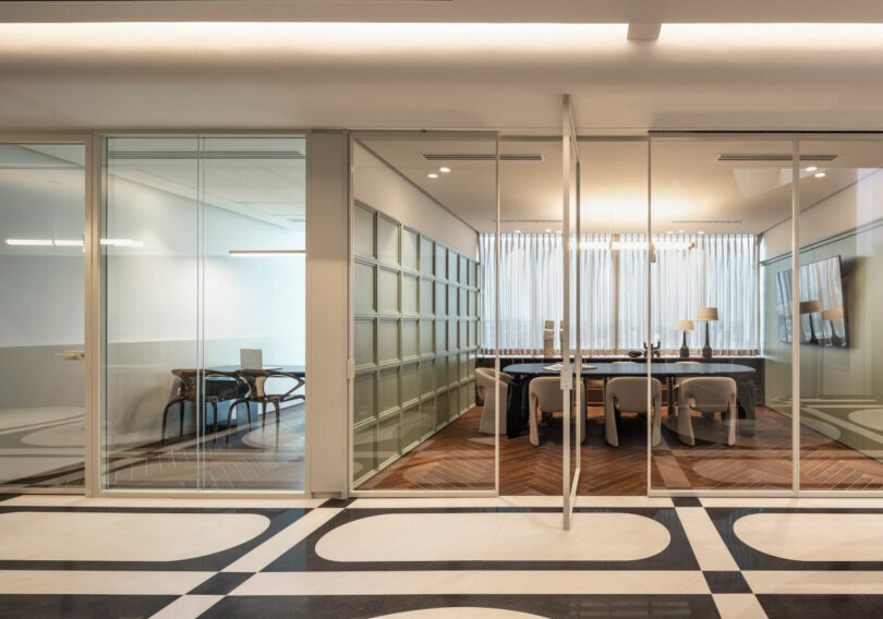 Modern office interior with glass walls, two meeting rooms furnished with tables and chairs, patterned floor, and natural light coming through sheer curtains.