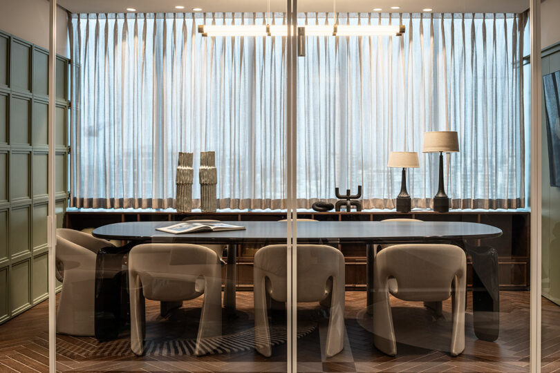 Modern conference room with a long table, six beige chairs, two table lamps, decorative objects, and sheer curtains covering large windows.