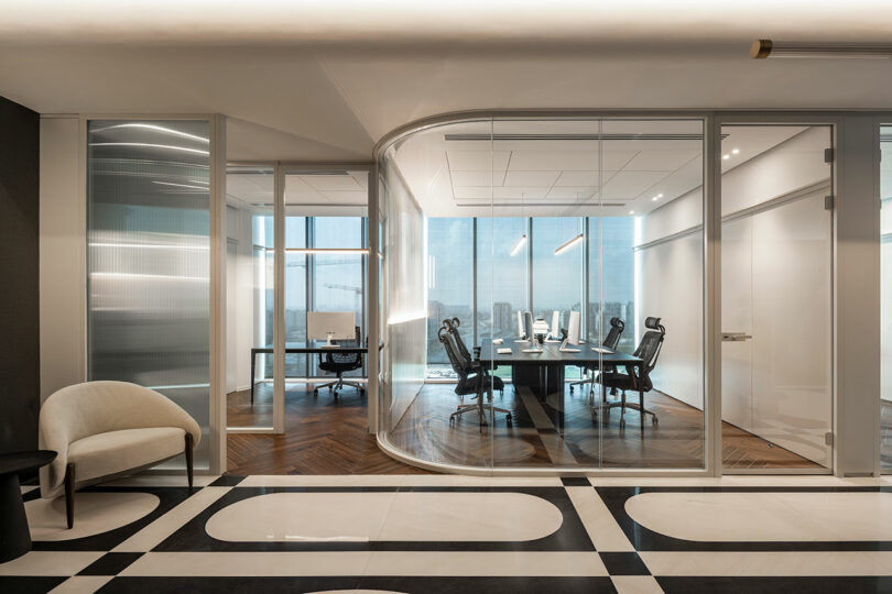 Modern office with glass-walled meeting room, long table, ergonomic chairs, wooden floor, and large windows allowing natural light; a single chair sits in the hallway.