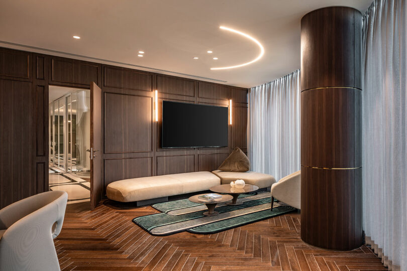 Modern living room with wood-paneled walls, a mounted TV, beige leather seating, round coffee table, patterned rug, large column, and floor-to-ceiling sheer curtains.