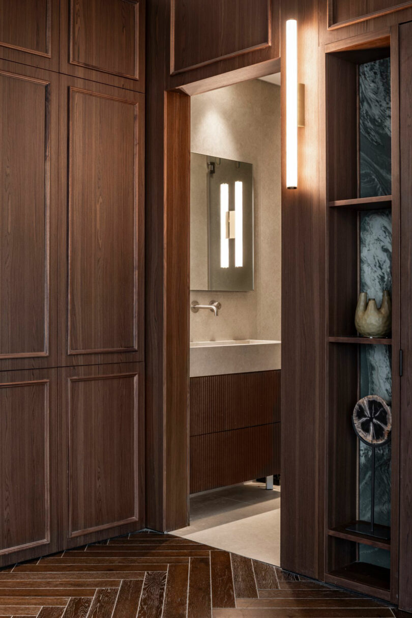 A wooden doorway leads to a modern bathroom with a wall-mounted sink, large mirror, and vertical light fixture; shelves and wood paneling are visible in the foreground.