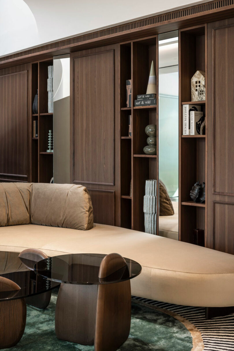 Modern living room with a curved beige sofa, dark glass coffee tables, and built-in wooden shelves featuring books, decor, and geometric accents.