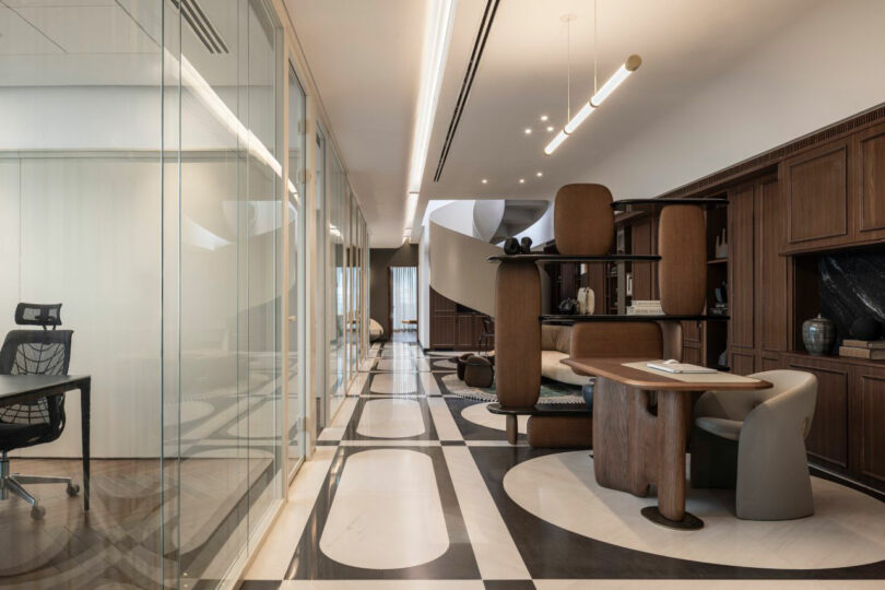 Modern office interior with glass-walled meeting room, wooden desk, shelves, and contemporary seating on a patterned tile floor.