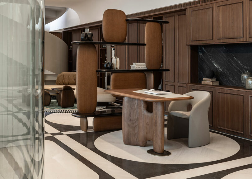 A modern office with wood paneling, a sculptural wooden desk, a curved beige chair, and open shelving dividing the space. Marble flooring features bold geometric patterns.