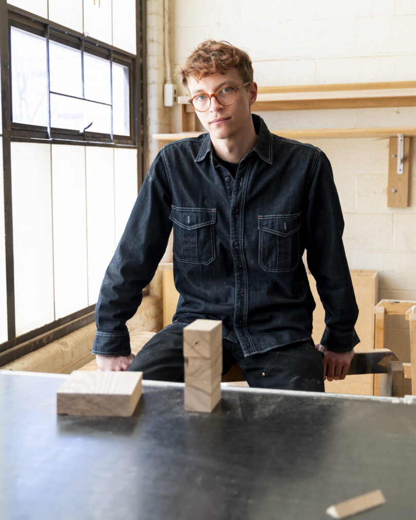 A person with short hair and glasses sits on a workbench in a woodworking shop, with wooden blocks and tools visible on the table and shelves