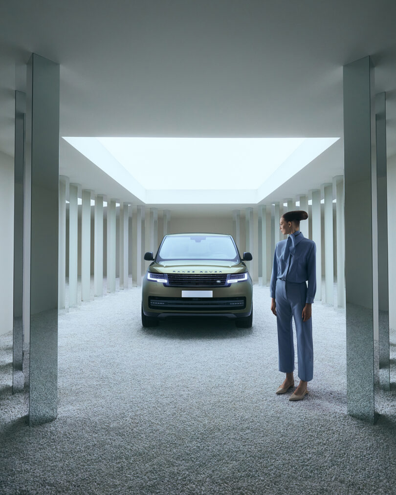 A woman in a blue suit stands in a modern, column-lined room, facing a parked SUV under a skylight.